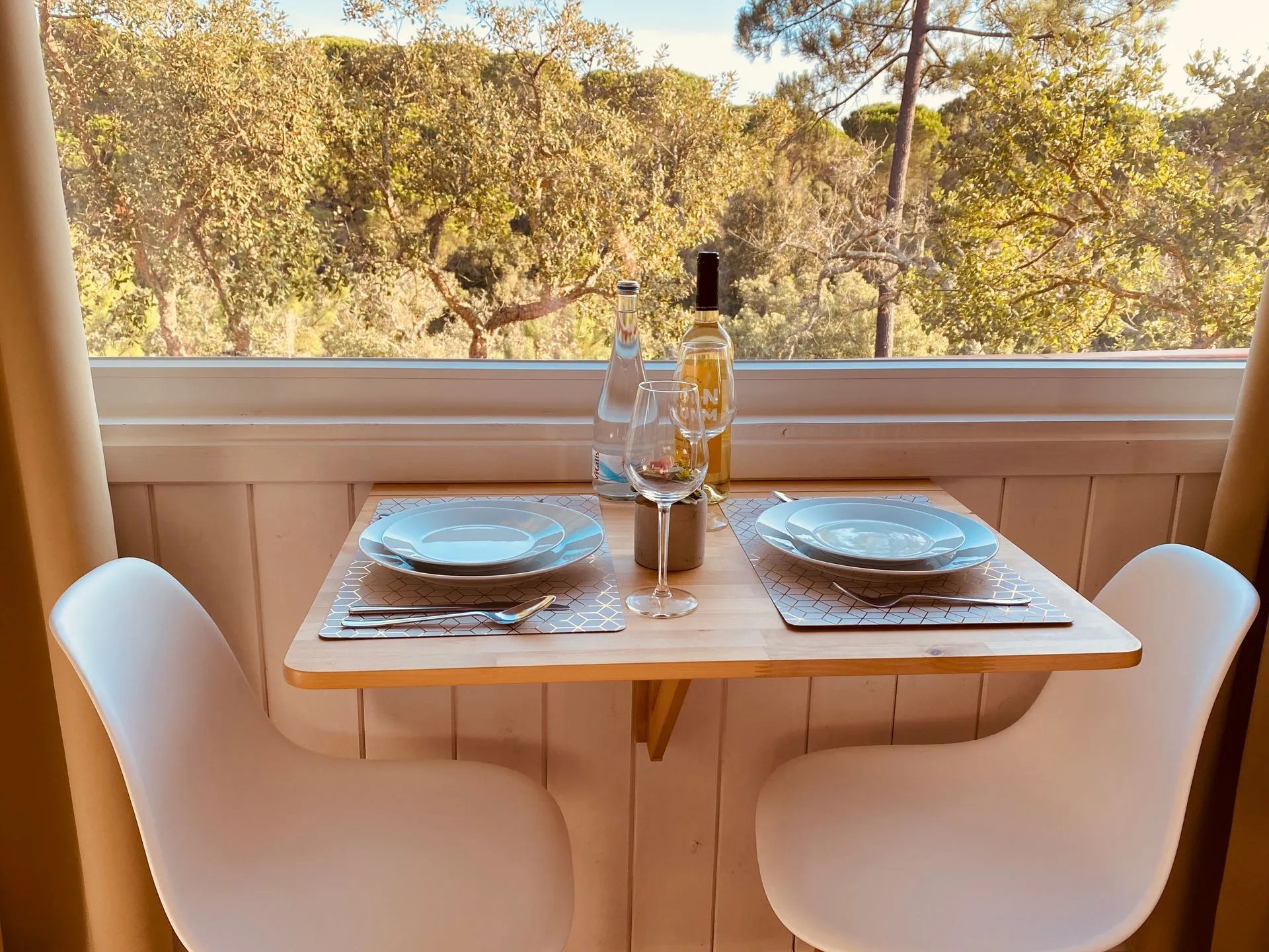 Una mesa de comedor con dos platos, una botella de vino y vasos, ubicada en un balcón con vista a árboles.
