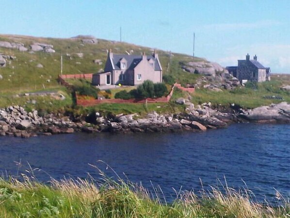A coastal house with a stone facade, a grassy hill in the background, and a rocky shoreline.