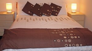 Uma cama bem arrumada com colcha estampada e almofadas, duas luminárias de cabeceira e uma mesinha de cabeceira de madeira com uma gaveta.
