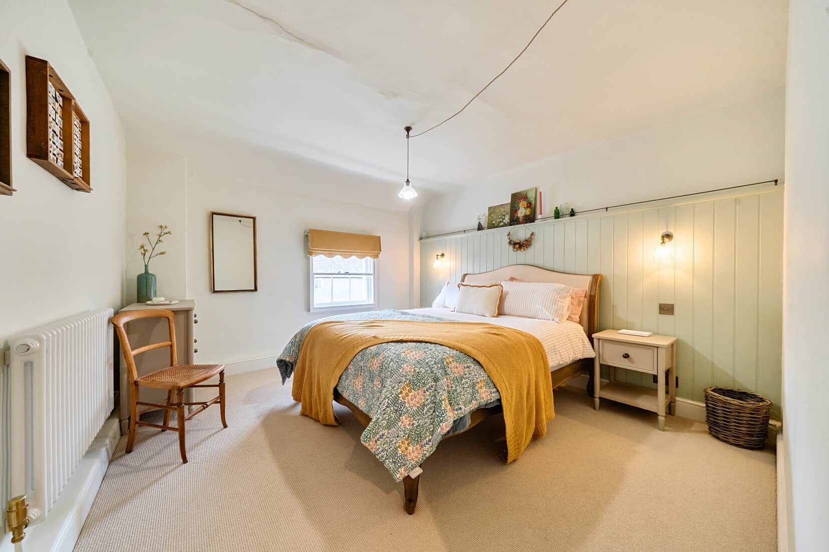 A bedroom with a bed, a chair, a nightstand, a radiator, a window with a blind, and a wall-mounted light fixture.