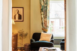 A room with a dark armchair, a radiator, a window with curtains, a framed picture on the wall, and a small table with books.