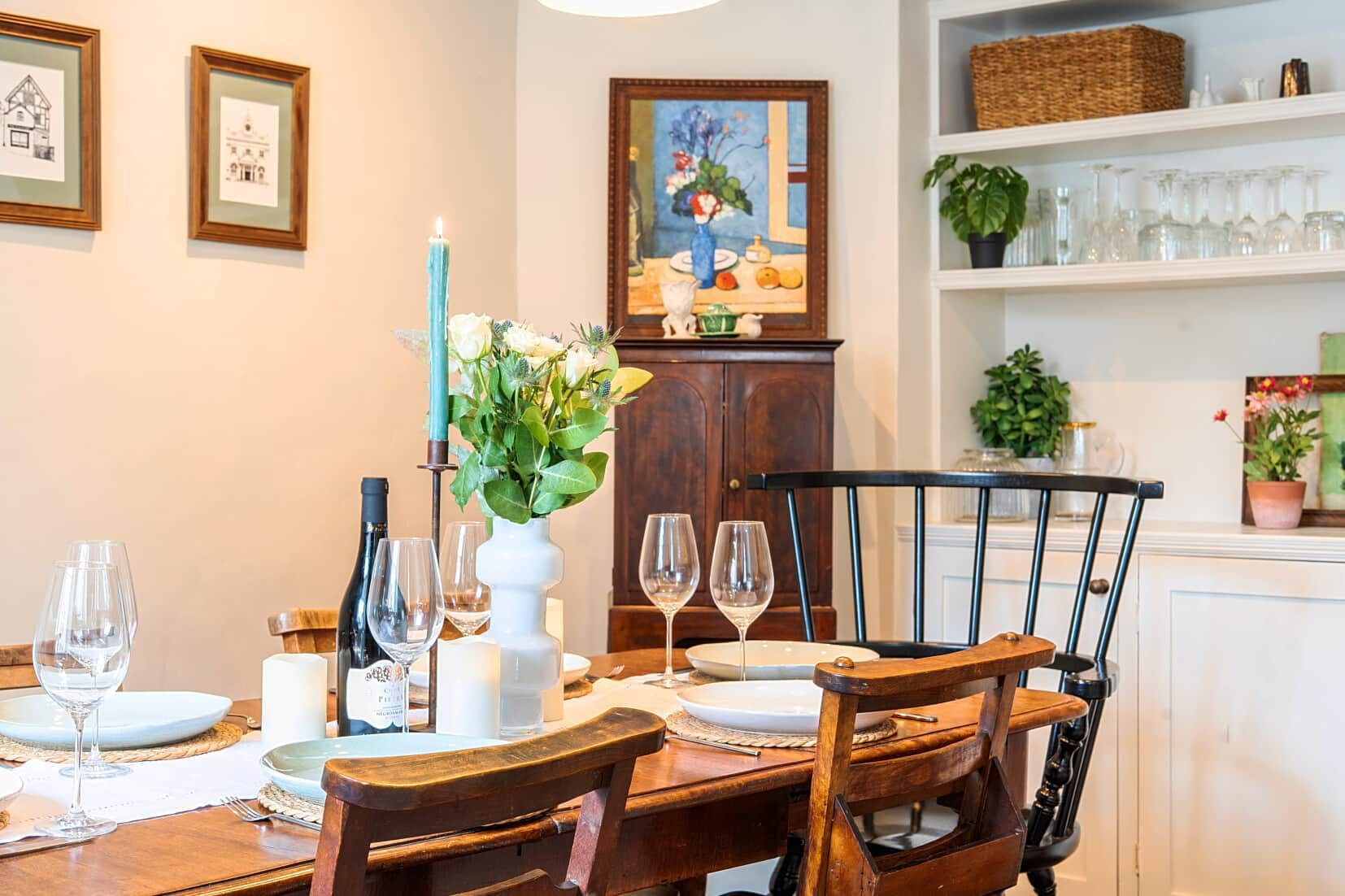 Mesa lista con copas de vino, platos y una vela. En la pared hay cuadros enmarcados y un mueble con un cuadro encima.