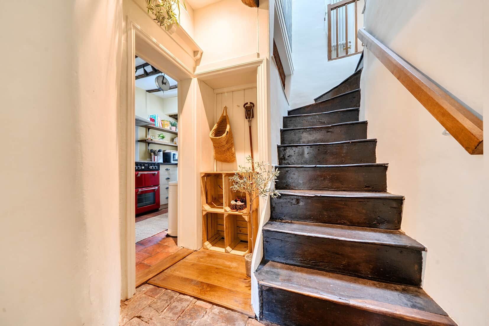 A wooden staircase leading to a second floor, a kitchen area with a red oven, and a basket hanging on the wall.