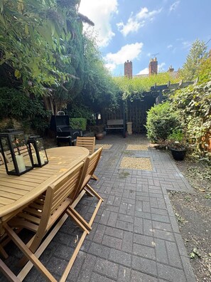 An outdoor patio with a wooden table and chairs, surrounded by greenery and a brick pathway. - Characterful Townhouse | Central 3 Bed + Parking (Ludlow)