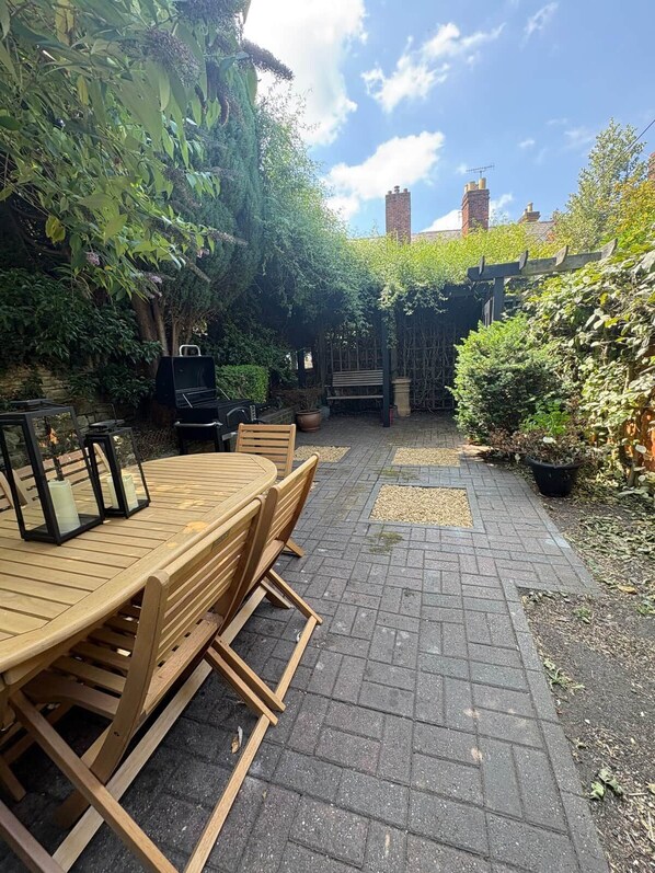 En udendørs terrasse med et træbord og stole, omgivet af grøn vegetation og en belagt sti.