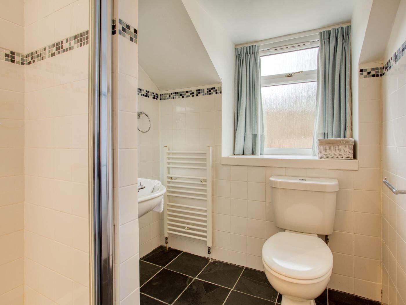 A bathroom with a white toilet, a white sink, a towel rack, a window with curtains, and a tiled floor.