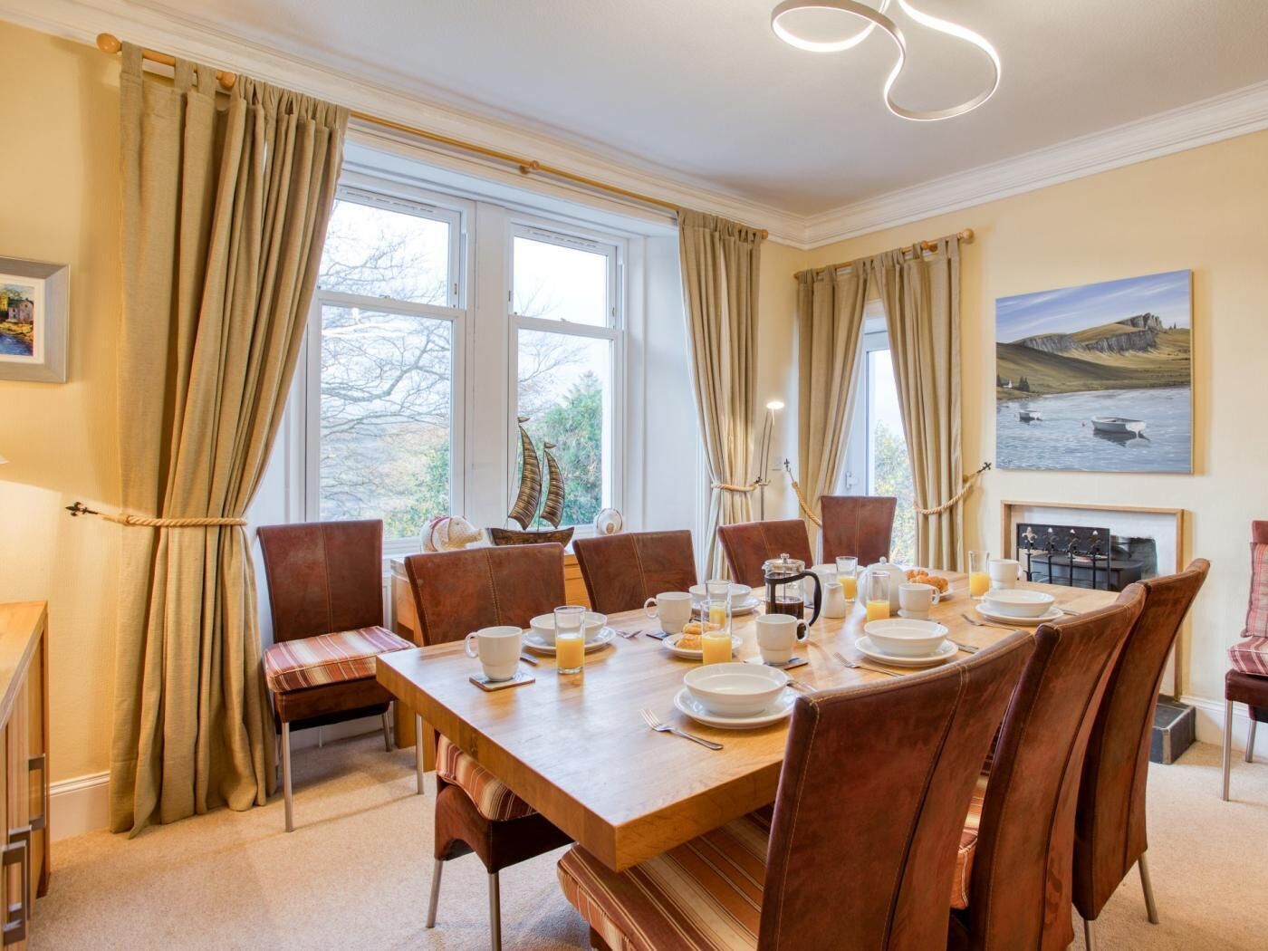 A dining room with a wooden table set for breakfast, large windows with curtains, and a painting on the wall.