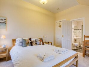 Une chambre à coucher avec un lit, des oreillers, une table de chevet avec une lampe, une porte donnant sur une salle de bain et une petite table avec un livre.