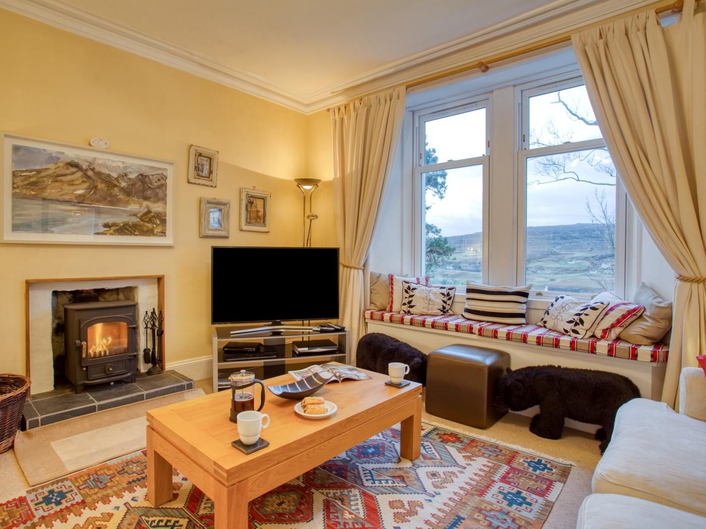 A cozy living room with a fireplace, a television, a sofa, a window seat, and a rug.
