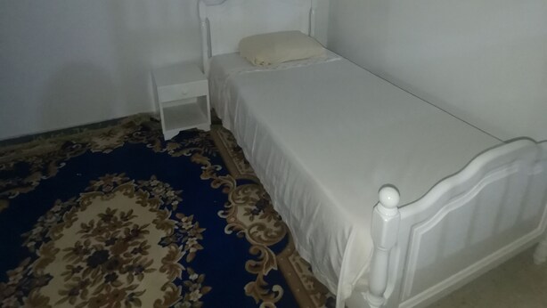Habitació petita amb un llit individual, un capçal blanc, una tauleta de nit i una catifes amb disseny.