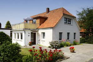 A two-story house with a red-tiled roof, a balcony, and a small garden with red flowers. - Holiday Home 'Ankerplatz' in Butjadingen – Comfort for up to 5 Guests (Butjadingen)