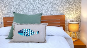 Un lit avec une tête de lit en bois, deux oreillers, une lampe et un mur orné d’un motif végétal.
