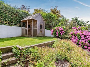 Ein hölzernes Gartenhaus mit Veranda, umgeben von einem gepflegten Garten mit verschiedenen Pflanzen und Blumen.