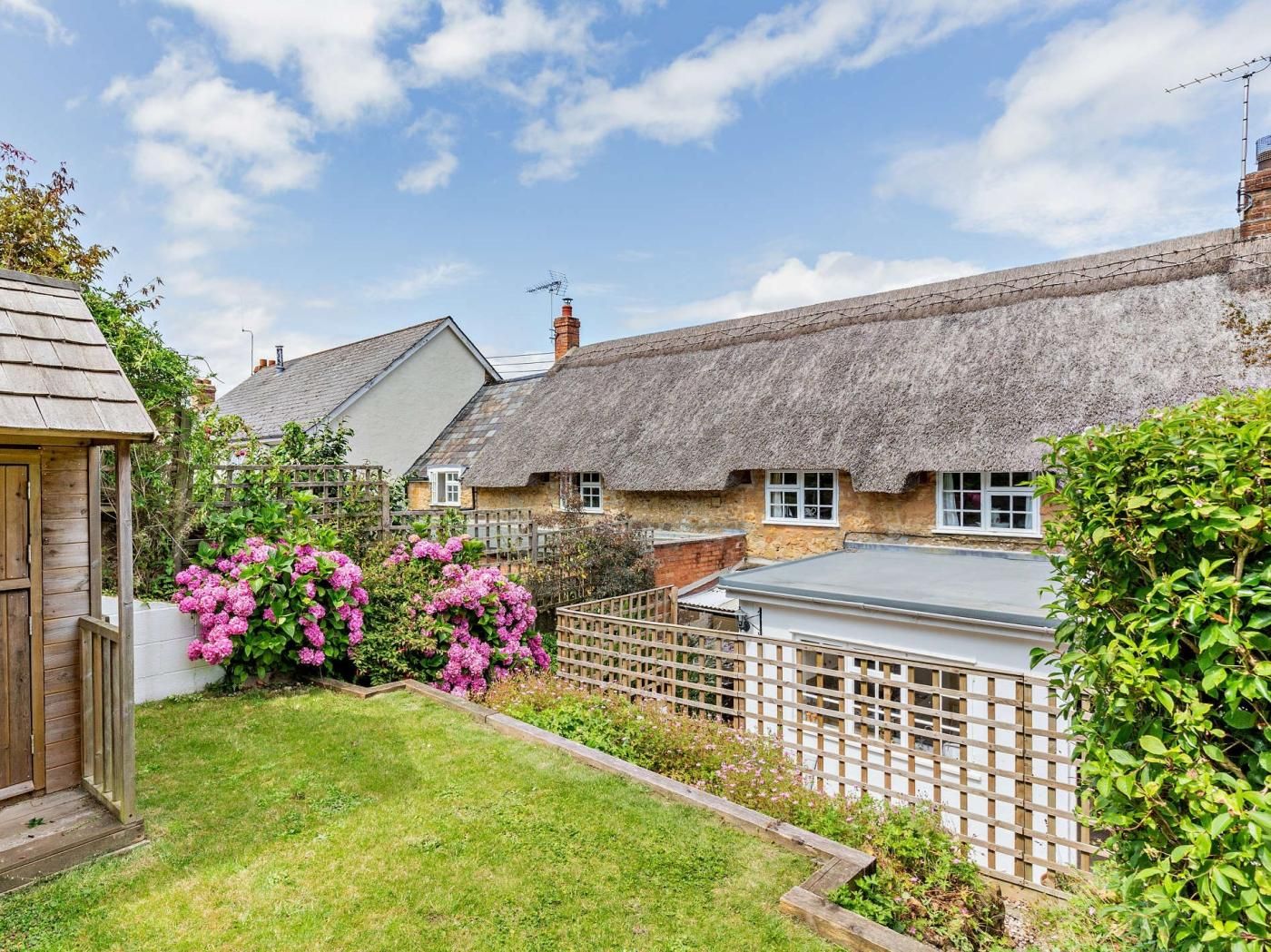A traditional thatched-roof house with a wooden fence and a garden with pink flowers.