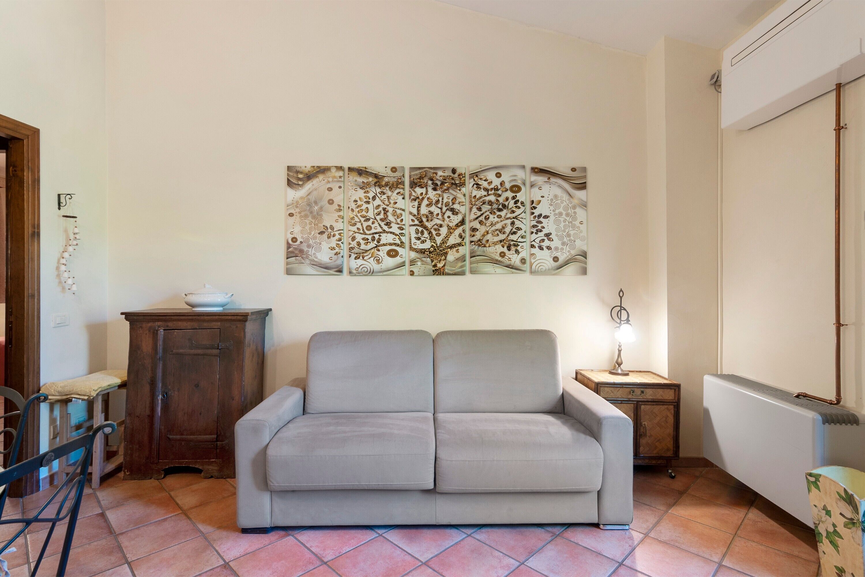Uma sala de estar com sofá, armário, mesa com abajur e uma obra de arte na parede.