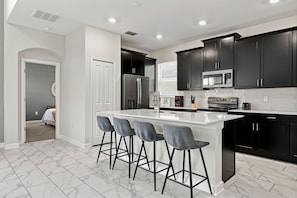 A modern kitchen with dark cabinets, a white island, and bar stools. - 5BR ChampionsGate Home w/ Pool & Theme Rooms (Kissimmee)
