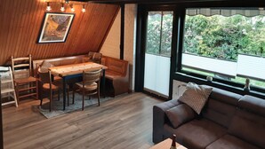 Een gezellige kamer met een houten eettafel, stoelen, een bank en een groot raam met uitzicht op groen.