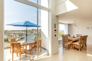 A modern dining area with a table and chairs, a view of a body of water, and a balcony with a blue umbrella. - 5 Bed in Fowey (oc-77657) (Polruan)