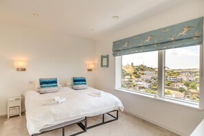 A bedroom with a bed, two pillows, a bedside table, a lamp, and a window with a view of a town. - 5 Bed in Fowey (oc-77657) (Polruan)