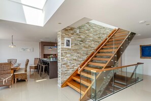 A modern interior with a glass staircase, a dining area with wicker furniture, and a kitchen in the background. - 5 Bed in Fowey (oc-77657) (Polruan)