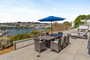 An outdoor patio with a table and chairs, a blue umbrella, and a view of a harbor with boats. - 5 Bed in Fowey (oc-77657) (Polruan)
