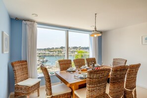 A dining area with wicker chairs and a wooden table set for six, overlooking a scenic view of a river and buildings. - 5 Bed in Fowey (oc-77657) (Polruan)