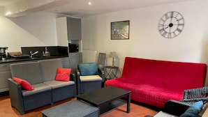 Ein Wohnbereich mit einem roten Sofa, zwei grauen Sofas, einem Couchtisch, Korbstühlen und einer Uhr, im Hintergrund ist eine Küche erkennbar.