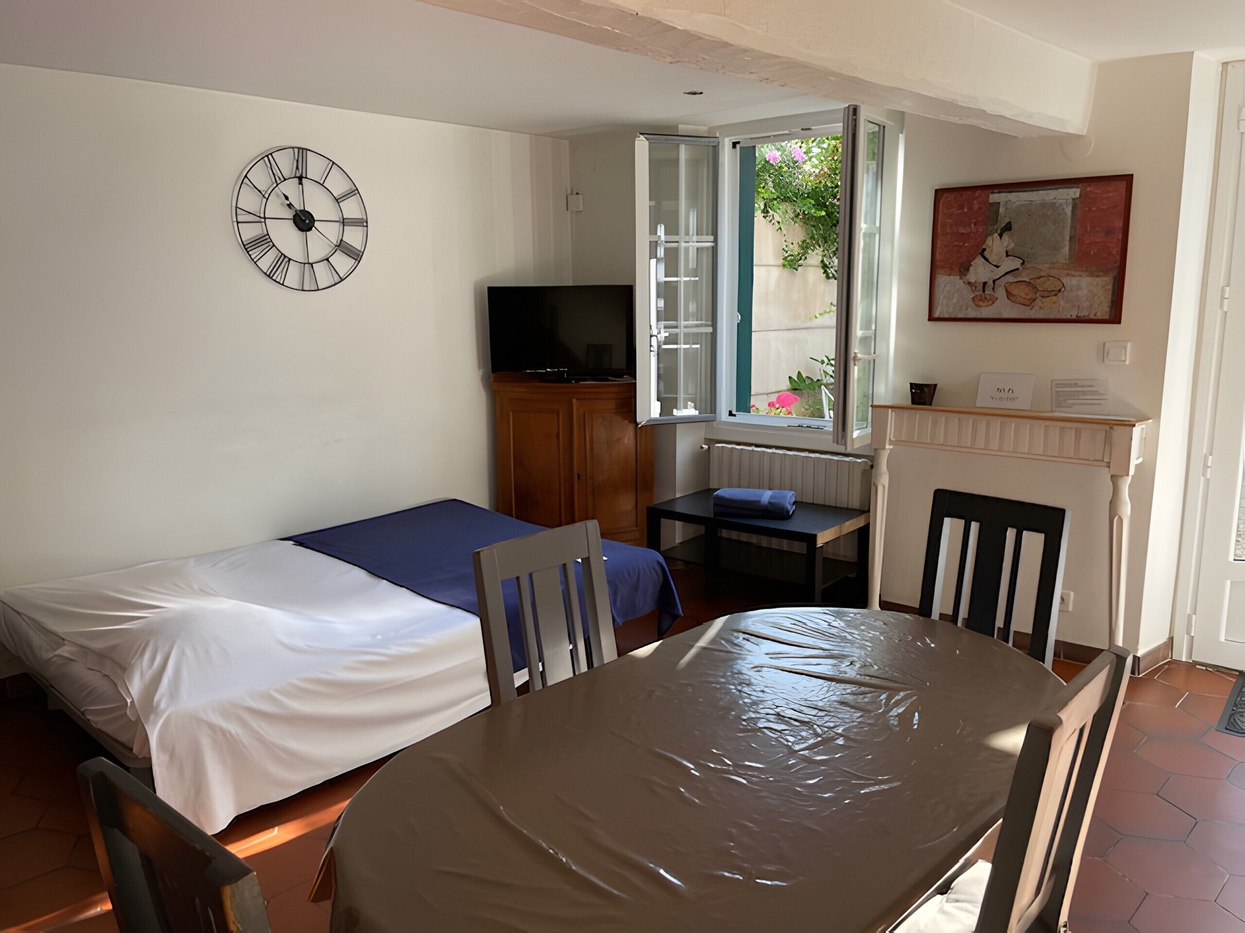 A room with a bed, a table with chairs, a TV, a window with a view, and a clock on the wall.