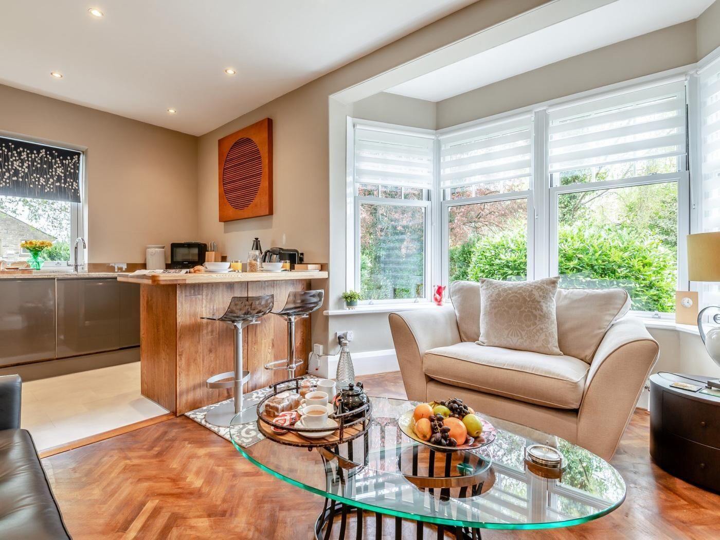 Eine moderne Küche mit Bar, ein Glastisch mit Früchten und Gebäck, eine Couch mit Kissen und große Fenster mit Blick auf Grünflächen.