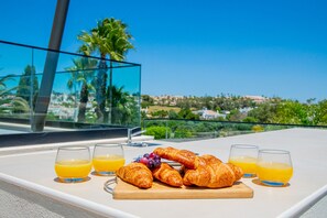En bakke med morgenmad, blandt andet croissanter, druer og appelsinjuice, på en balkon med udsigt over et smukt landskab.