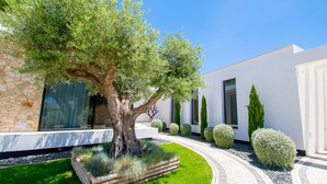 A modern building with a stone facade, a large tree in the center, and a well-maintained garden with hedges and a pathway. - Villa Cactus in Carvoeiro (Lagoa)