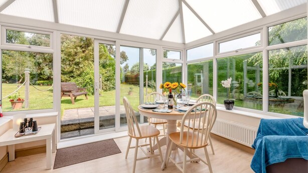 Een oranjerie met een eettafel, stoelen, een bankje en uitzicht op een tuin en vijver.