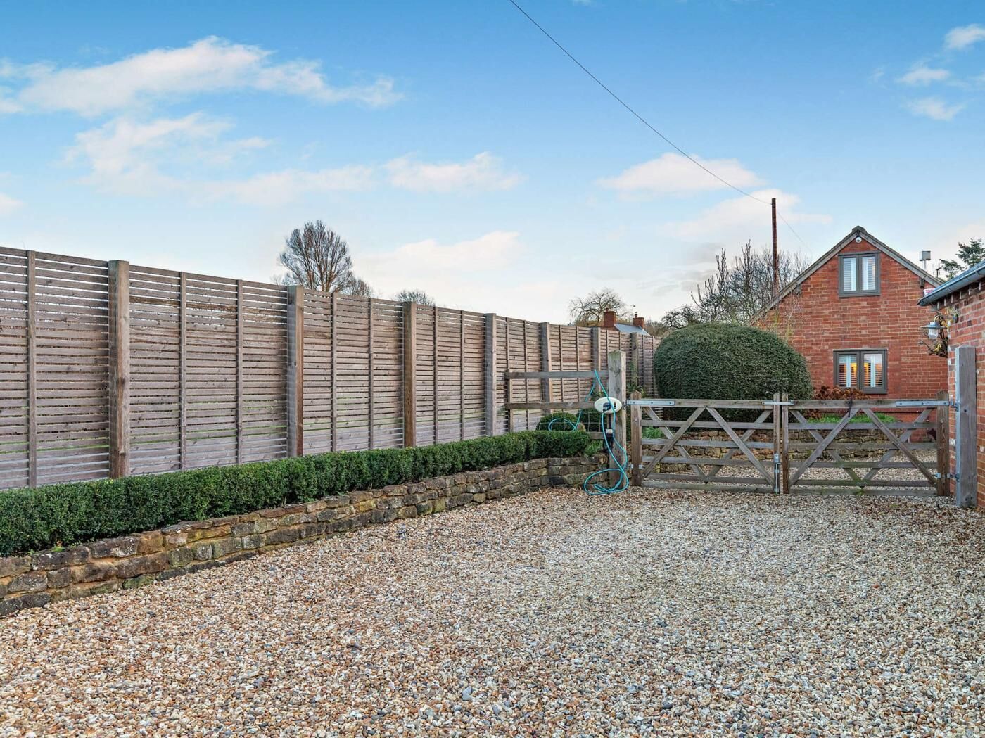 A gravel driveway leading to a wooden gate, a stone wall, and a brick house with a red roof.