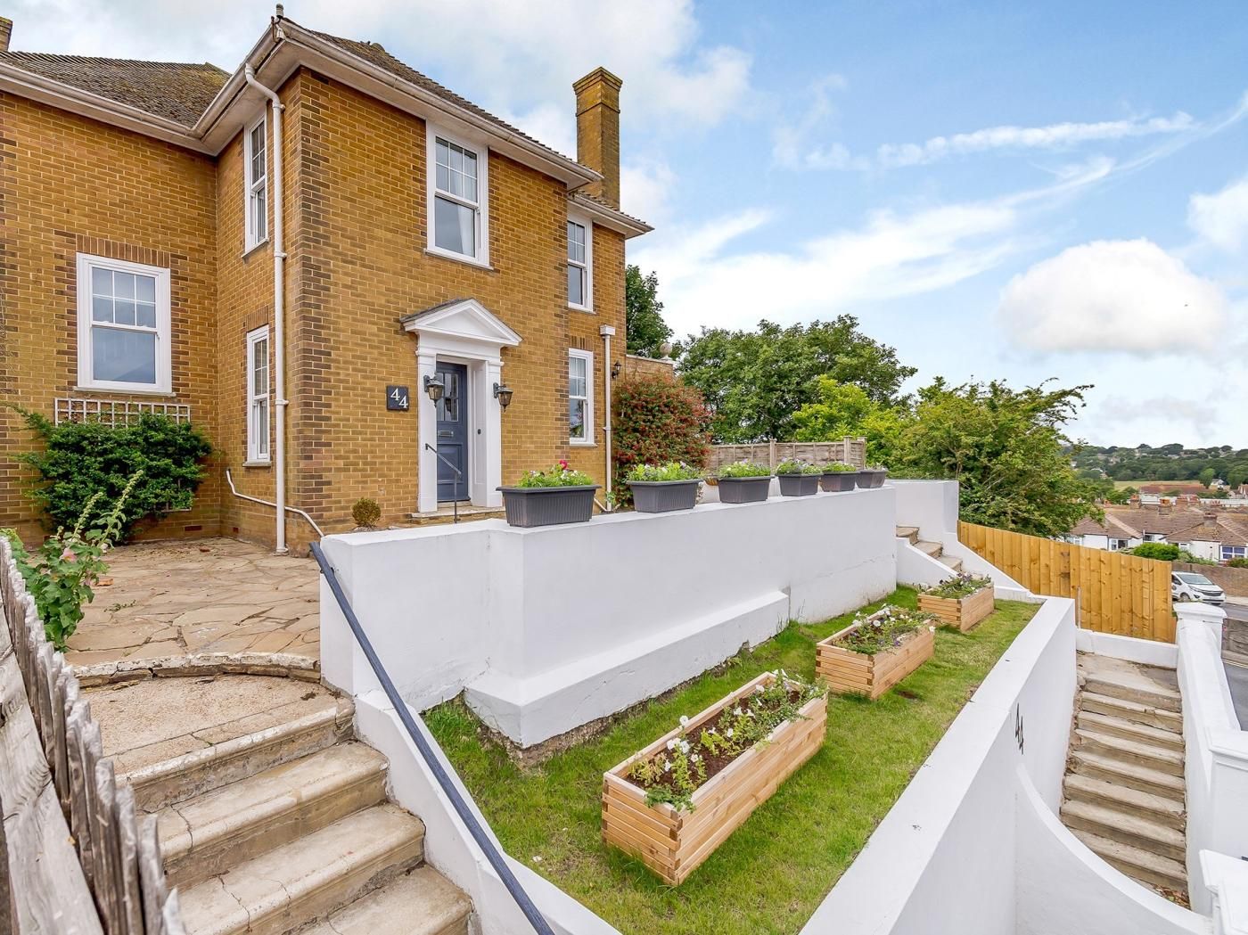 A two-story brick house with a front door, a small front garden with raised beds, and a white staircase leading to the entrance.