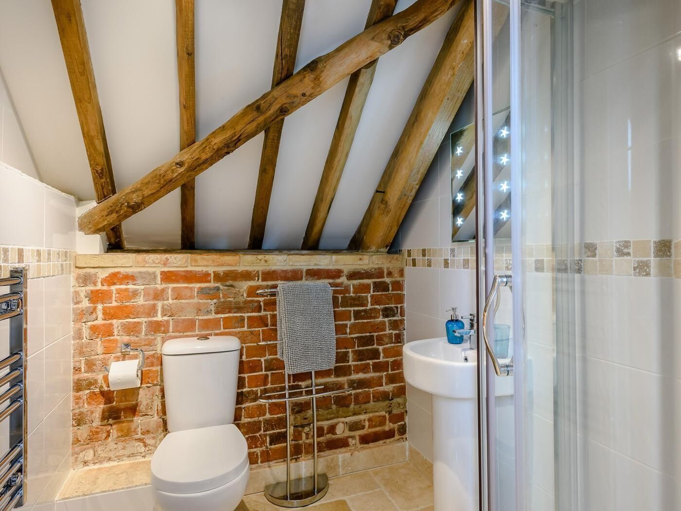 Un baño con pared de ladrillo, vigas de madera, inodoro, lavabo y cabina de ducha de vidrio.