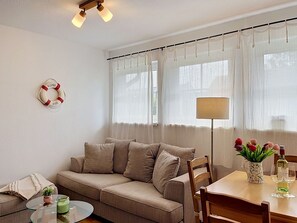 A living room with a sofa, a glass coffee table, a dining table with chairs, a lamp, and a window with sheer curtains. - Nice apartment for 4 guests with WIFI, TV and terrace (Wolgast)