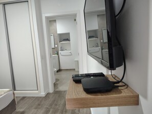 Ein modernes Hotelzimmer mit einem Nachttisch aus Holz, einem an der Wand montierten Fernseher und einem durchgegangenen Tür erkennbaren Badezimmer.