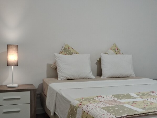 Ein Schlafzimmer mit einem Bett, zwei Kissen, einer Nachttischlampe, einem Nachttisch mit Schubladen und einer Steudecke.