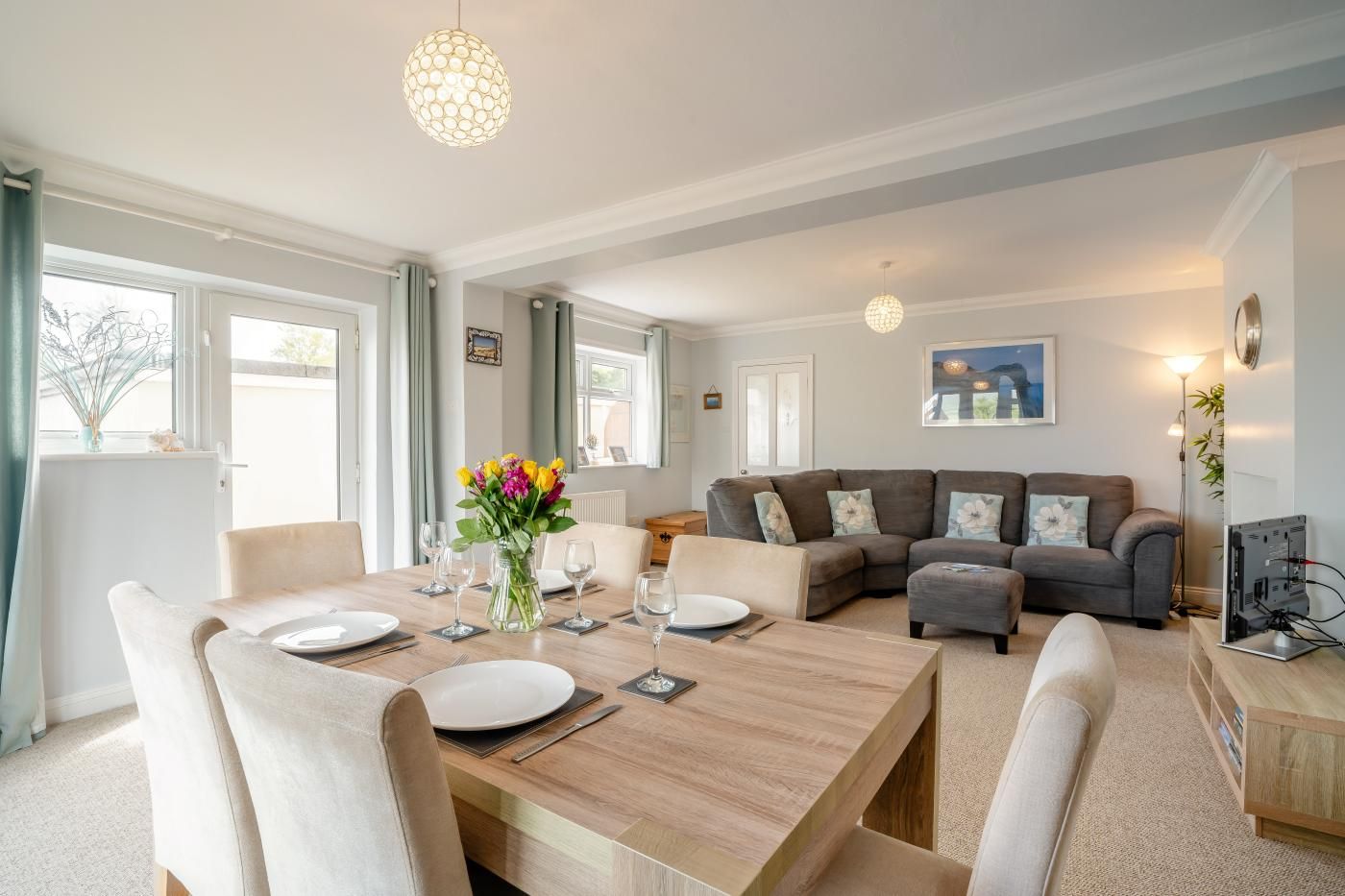 A modern living room with a dining area, a grey sofa set, a chandelier, and a painting on the wall.
