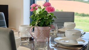 Uma mesa de jantar posta com pratos, tigelas e copos brancos, um vaso de flores cor-de-rosa com rosas, e vista de um campo pela janela.