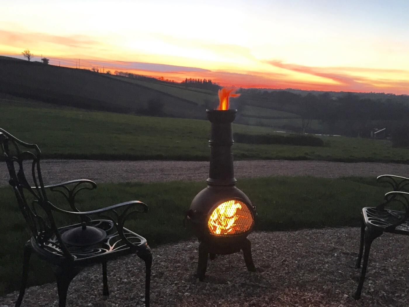 Un fogón de leña con fuego encendido, dos sillas de metal y un camino de grava.
