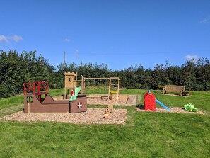 Ein Spielplatz mit verschiedenen Spielgeräten, darunter eine Rutsche, Schaukeln und Kletterbereiche, inmitten einer grünen Wiese. Im Hintergrund sieht man eine Holzbank und eine Baumreihe.