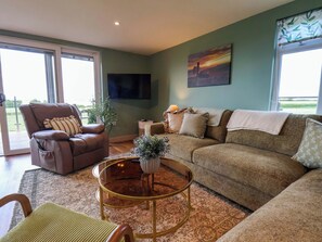 A living room with a brown leather sofa, a glass coffee table, a brown armchair, a TV mounted on the wall, and a window with a view. - Freedom Lodge @ Summerfield Farm (Whitby)