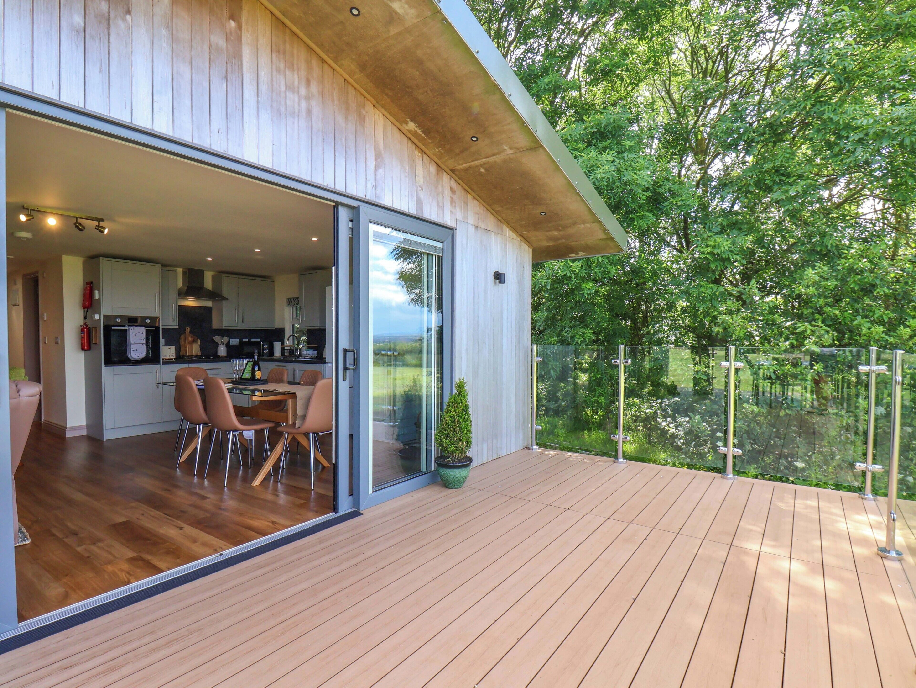 Une maison moderne dotée d’une terrasse en bois, d’une porte vitrée donnant sur un coin repas et offrant une vue sur la verdure.