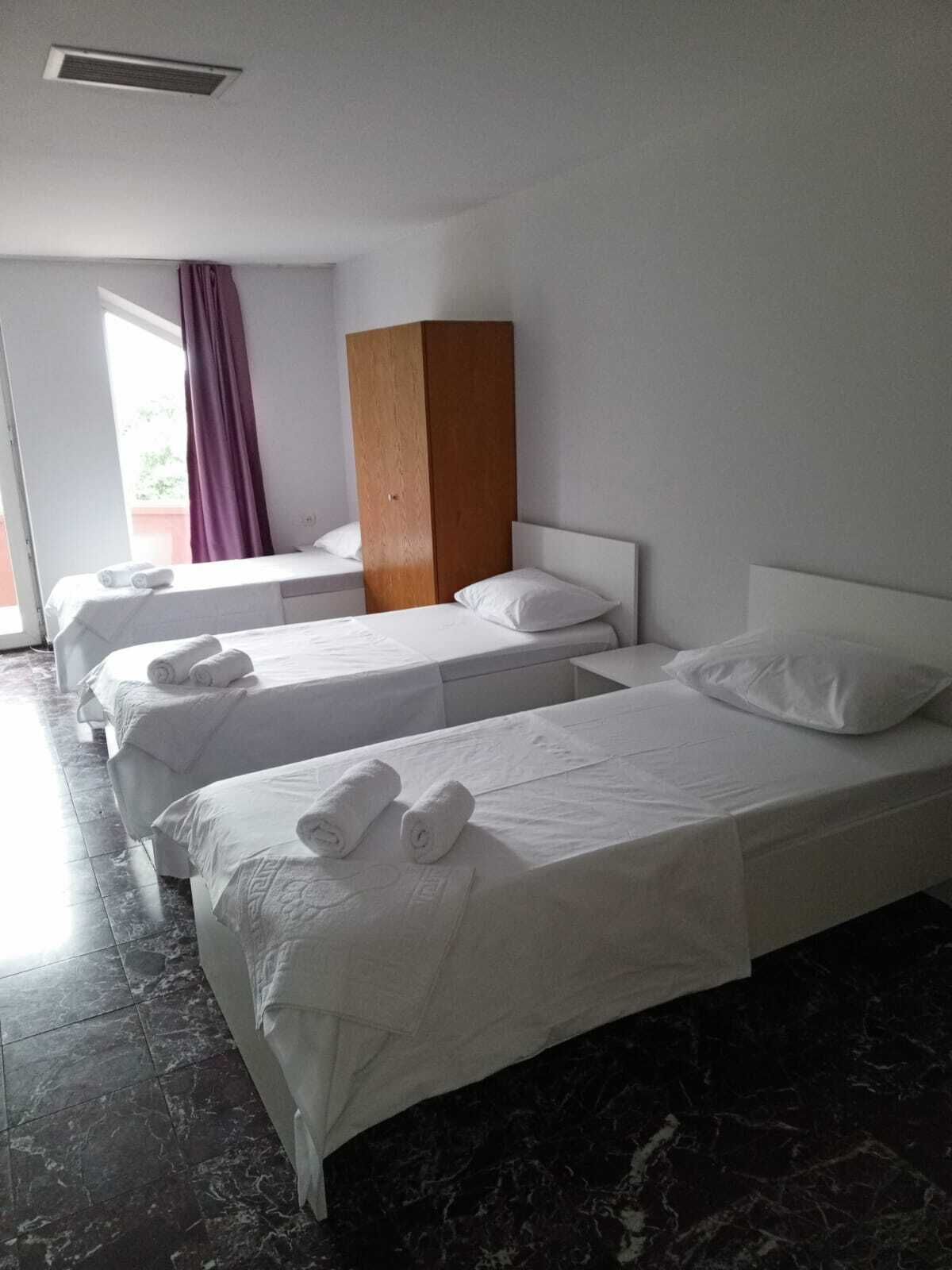 Habitació d'hotel amb tres llits individuals, un armari de fusta, una finestra amb cortines morades i un paviment de marbre.