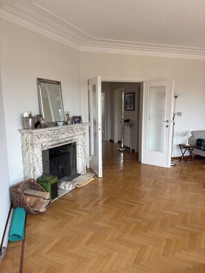A spacious living room with a marble fireplace, a large mirror, and a herringbone wooden floor.