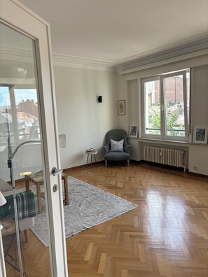 Una habitación amplia con suelo de parquet de madera, un sillón gris, una mesita auxiliar y un radiador.