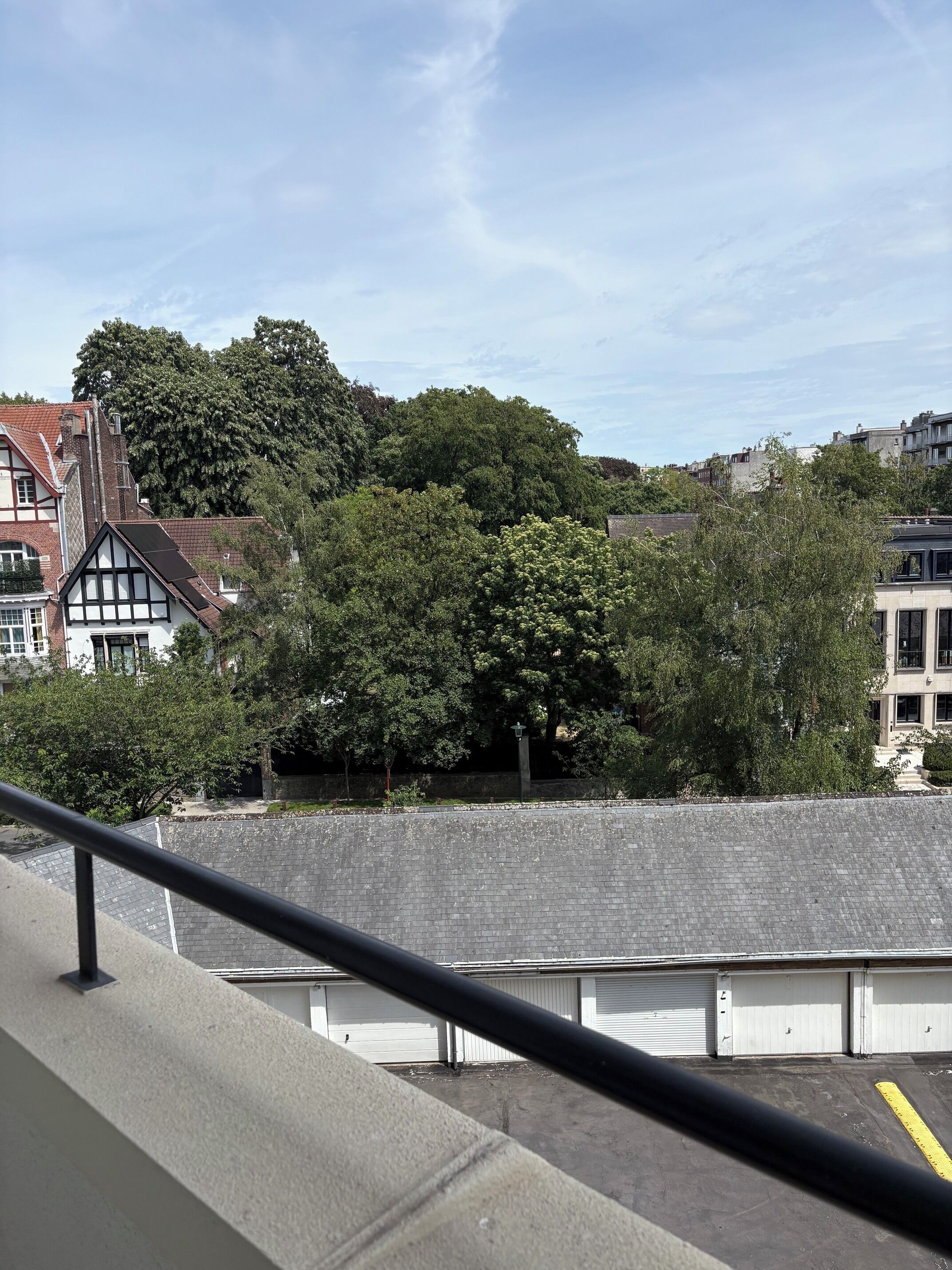 Vista de un edificio con balcón, zona pavimentada y un gran árbol al fondo.