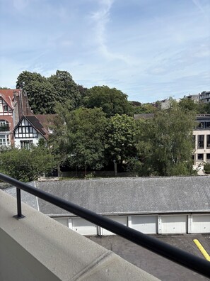 A view of a building with a balcony, a paved area, and a large tree in the background.