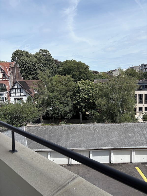 Vista de un edificio con balcón, zona pavimentada y un gran árbol al fondo.